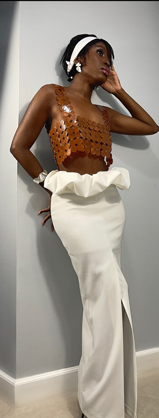 front view of model wearing a white long skirt with kick pleat slit in the center front of the gown. Skirt features a scrunchy puff that slightly dips over the top hem. Mimicking the bloom of a flower opening up. There is a zipper at the center back. The skirt is design with a slim fit similar to a long pencil skirt. Skirt is floor length.