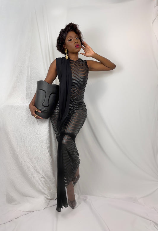 Front view of a black sheer mesh gown with a faux leather wave pattern that repeats all over the body. Garment features a black sheer drape that wraps from shoulder to a bow around the legs and a delicate and water like way. Garment is sleeveless and has a invisible zipper in the back. 