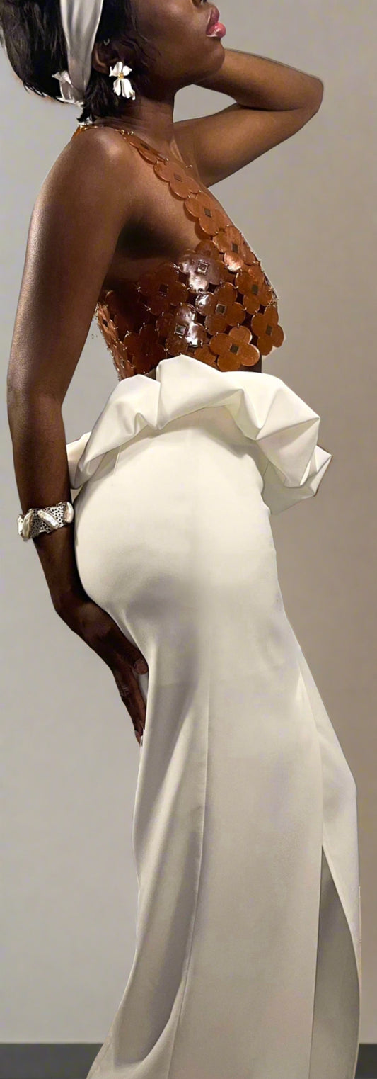 Side view of model wearing a white long skirt with kick pleat slit in the center front of the gown. Skirt features a scrunchy puff that slightly dips over the top hem. Mimicking the bloom of a flower opening up. There is a zipper at the center back. The skirt is design with a slim fit similar to a long pencil skirt. Skirt is floor length.