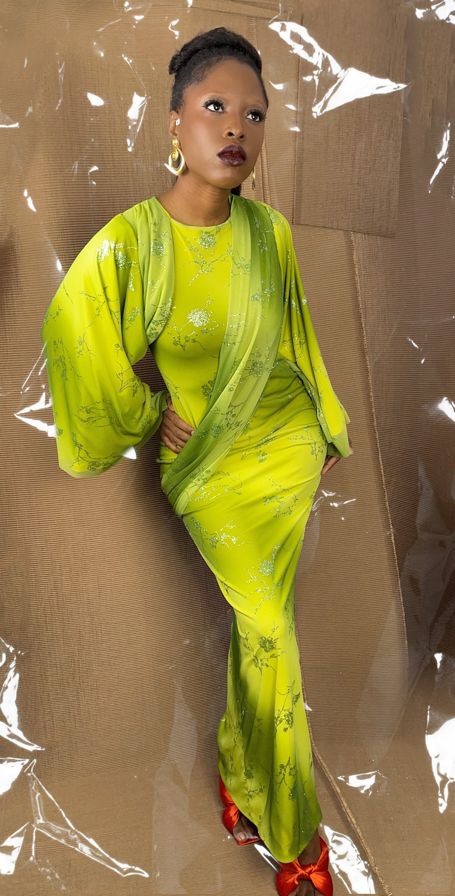 Front view of our modest flowy drape dress. The dress is made in a ombre green and yellow fabric with images of flowers in matching glitter. The dress features a sash like detail that wraps across the body from the front to the back. The sleeves features a asymmetric hem and bellow out to create more gathers. The dress is maxi length with a small slit on one side.