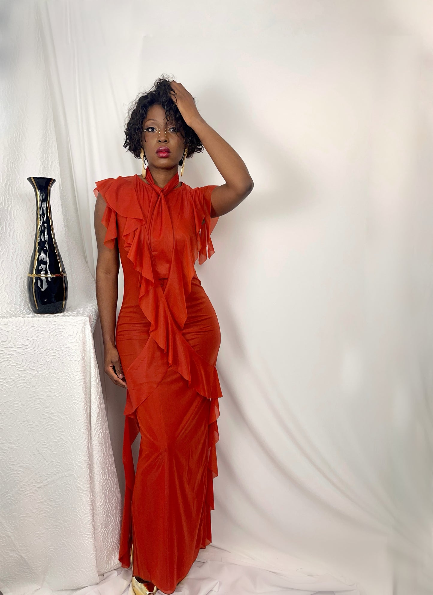 Front view of model wearing a full length dress in sunset orange. Flounces crisscross the front of the body then cascading down both sides of the dress. This garment is both elegant and flirty, with a modern twist with matching slim fit neck tie.