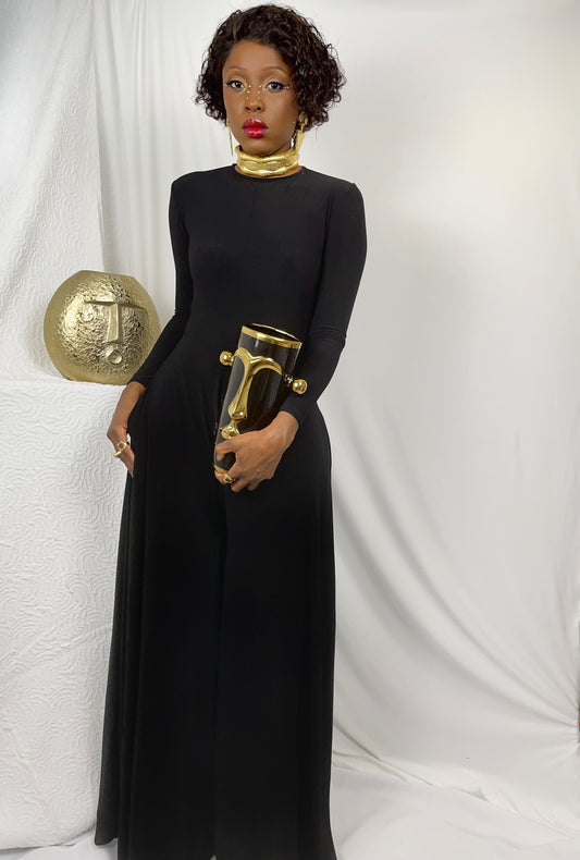 Front view of model wearing a slinky all black knit jumpsuit with flared wide leg pants. Garment features a snug comfy fit across the body then flaring out to a faux like skirt when standing still. Garment is made in a spandex knit for ease with inseam pockets.