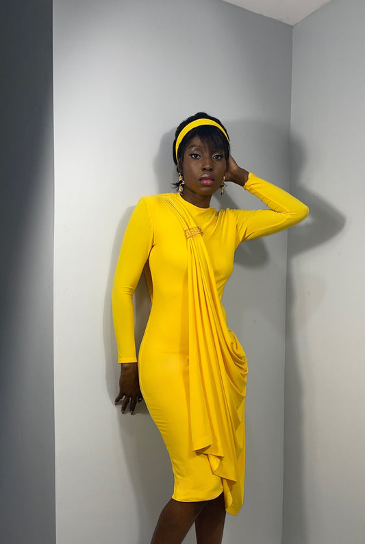 Model is posing in front of a wall wearing a yellow knit mid length dress. Dress features a drape cascades similar to a waterfall, and broach that was 3d printed to resemble honeycombs.  Great sophisticated summer look inspired by the honey bee. 