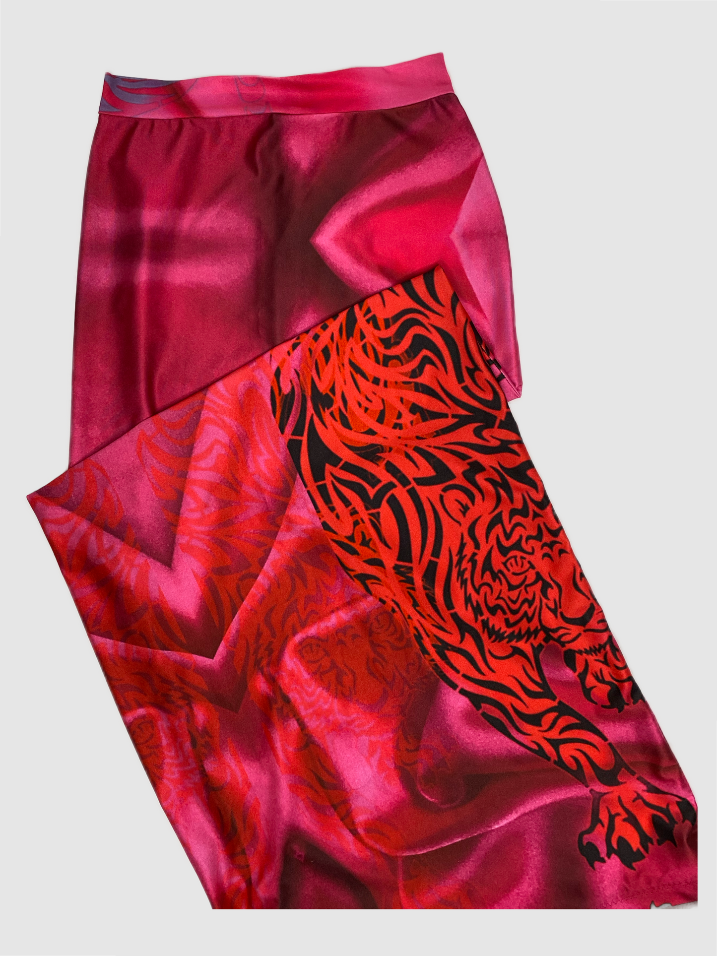 This floor length maxi is decorated in a custom print. The print ranges in color from light pink to fuchsia. There are images of tigers in a trio formation and geometric shapes in the background. The pattern is more large in scale. The tiger appear in and out of focus from a very light red, red and black and a light blue. The skirt has one side slit with black zipper. The slit can be worn open or closed. There is stretch and was crafted on a soft sheen Lycra material. 